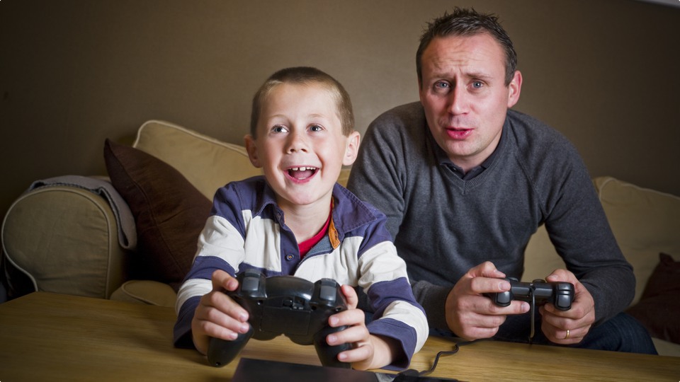 Glad man och pojke sitter i soffa och spelar dataspel.